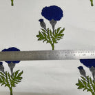 White fabric with a repeating pattern of dark blue flowers and green leaves, shown with a metal ruler across the center for scale.