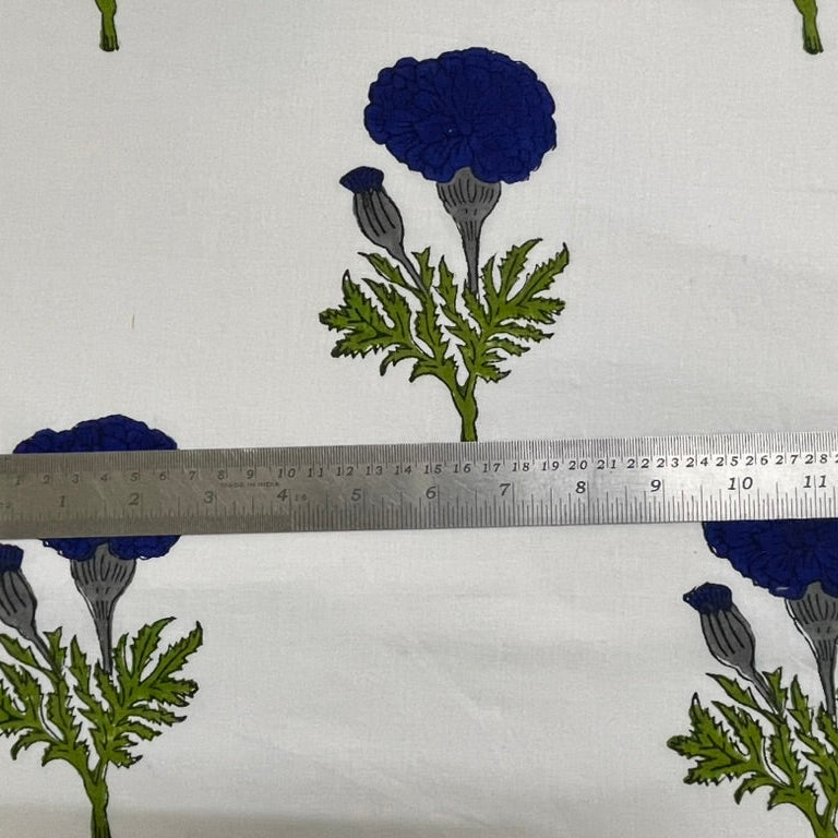 White fabric with a repeating pattern of dark blue flowers and green leaves, shown with a metal ruler across the center for scale.