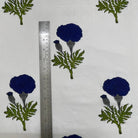 White fabric with a repeating block-print pattern of dark blue flowers and green leaves, shown next to a metal ruler for scale.