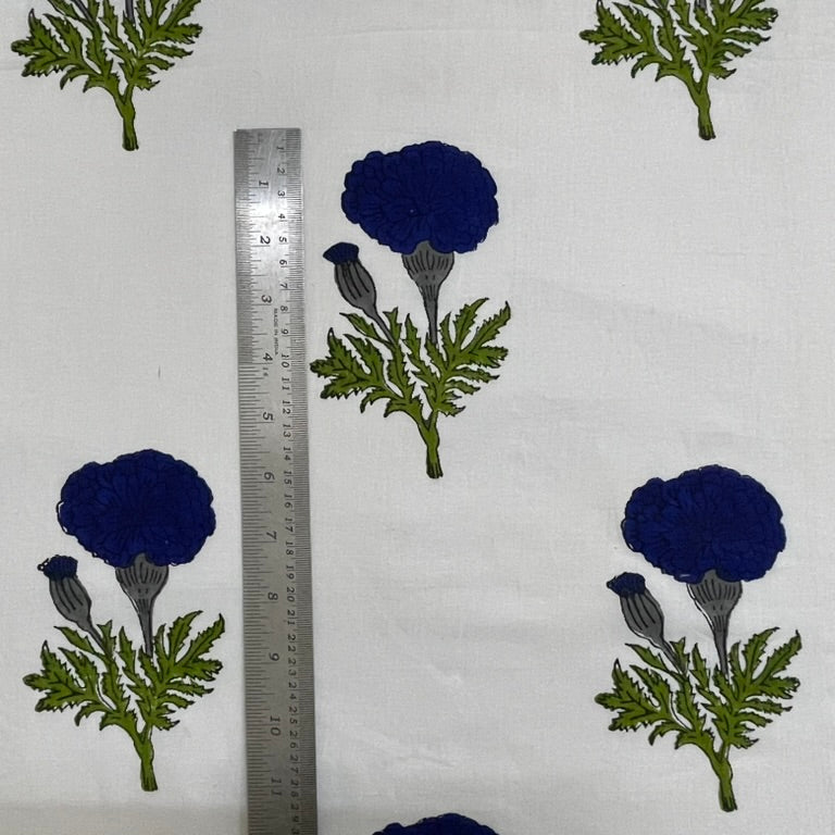White fabric with a repeating block-print pattern of dark blue flowers and green leaves, shown next to a metal ruler for scale.