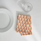 Napkin with white floral pattern on orange background next to a white plate and glass on a white surface